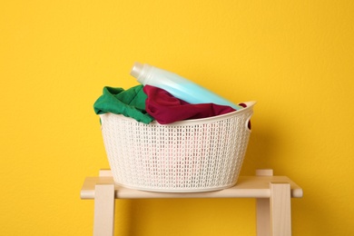 Laundry basket with dirty clothes and detergent on wooden stool against yellow background