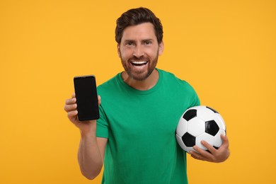 Photo of Happy sports fan with soccer ball and smartphone on orange background
