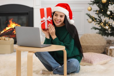 Celebrating Christmas online with exchanged by mail presents. Smiling woman in Santa hat with gift box during video call on laptop at home