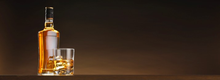 Image of Whiskey and ice cubes in glass and bottle on table against brown background, space for text. Banner design