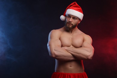 Attractive young man with muscular body in Santa hat on black background, space for text