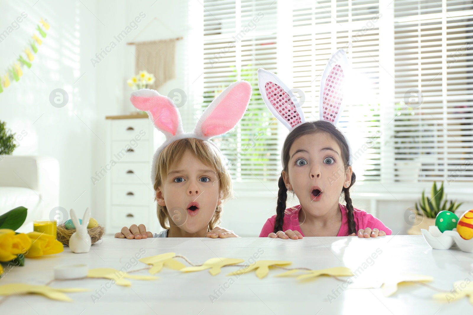 Photo of Emotional children wearing bunny ears headbands at table with Easter eggs, indoors