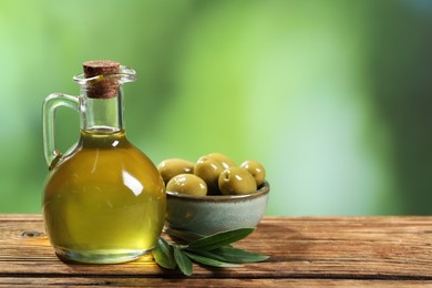 Photo of Jug of cooking oil, olives and green leaves on wooden table against blurred background. Space for text
