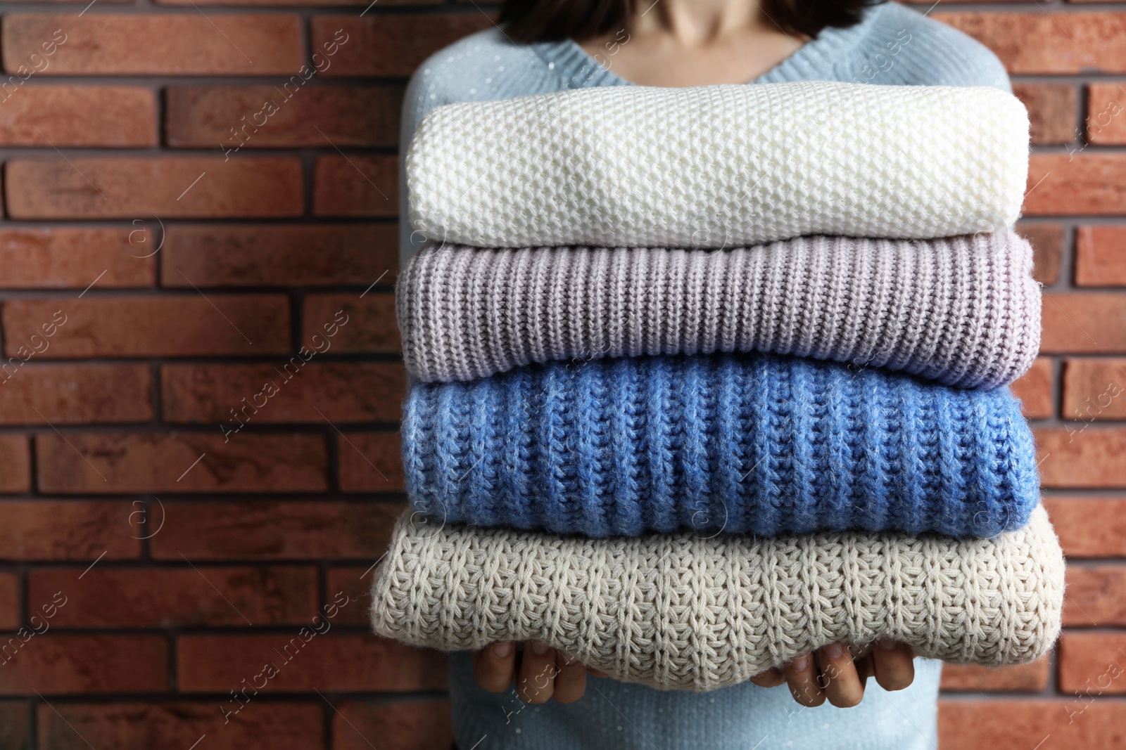 Photo of Woman holding pile of winter sweaters near brick wall, closeup view