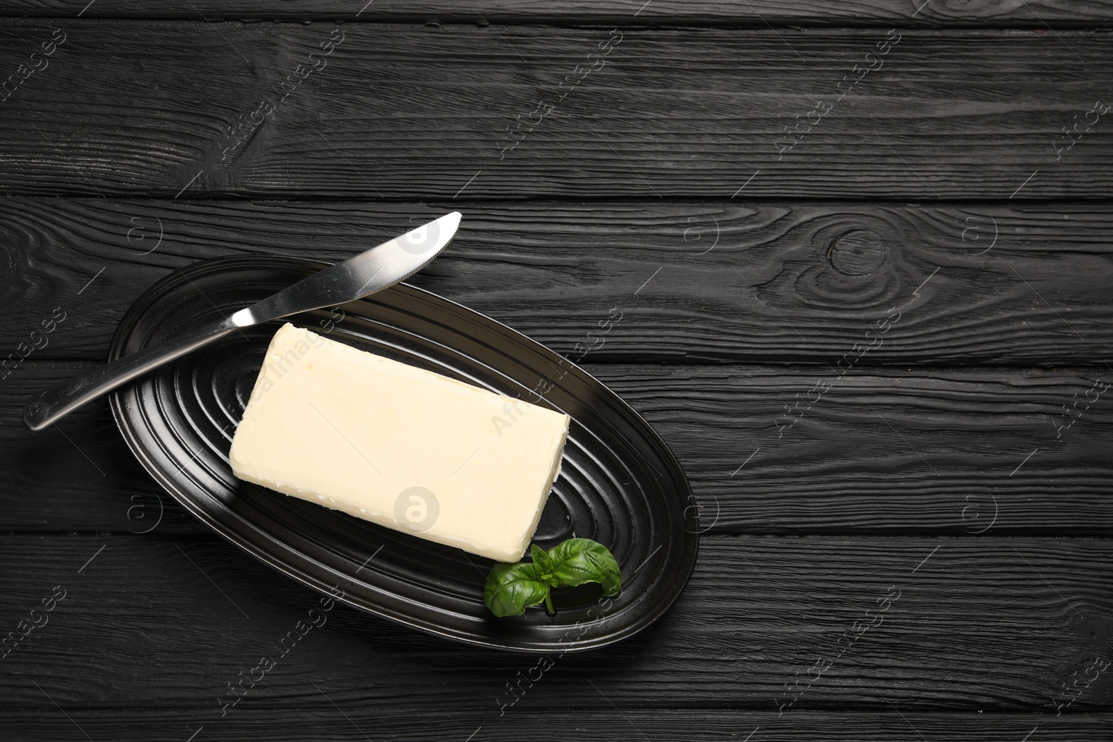 Photo of Block of tasty butter with basil and knife on black wooden table, top view. Space for text