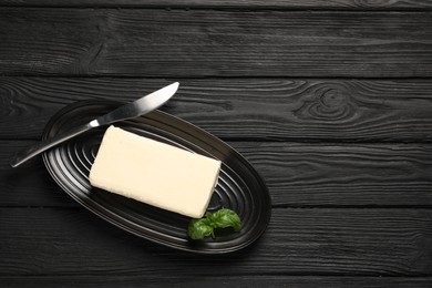 Block of tasty butter with basil and knife on black wooden table, top view. Space for text