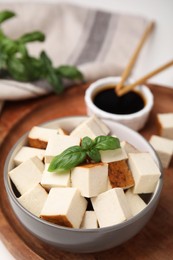 Photo of Bowl of smoked tofu cubes, soy sauce and basil on wooden tray, closeup