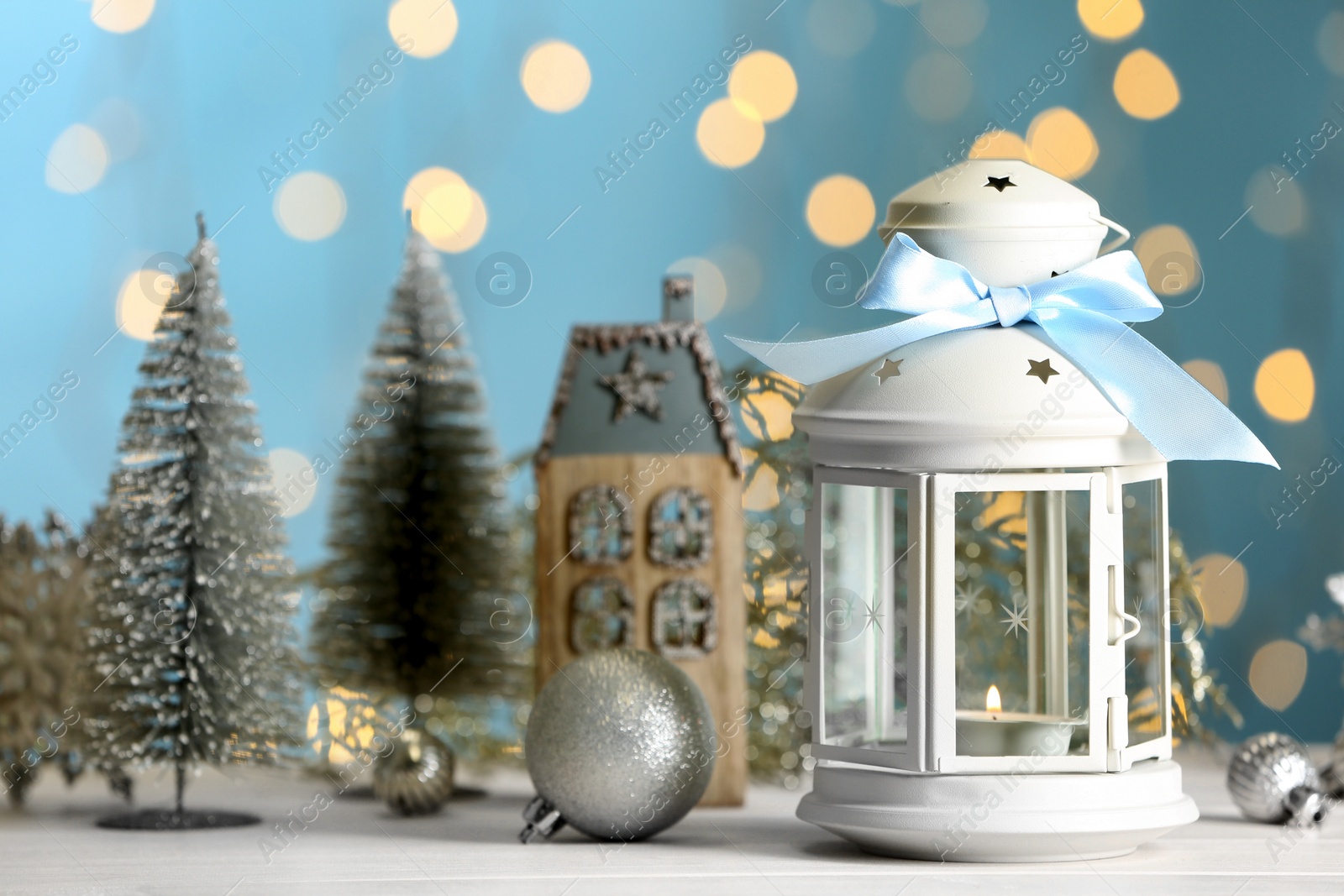 Photo of Christmas lantern with burning candle and festive decor on white table against blurred lights