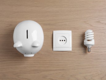 Photo of Piggy bank, power socket and fluorescent light bulb on wooden table, flat lay. Energy saving concept