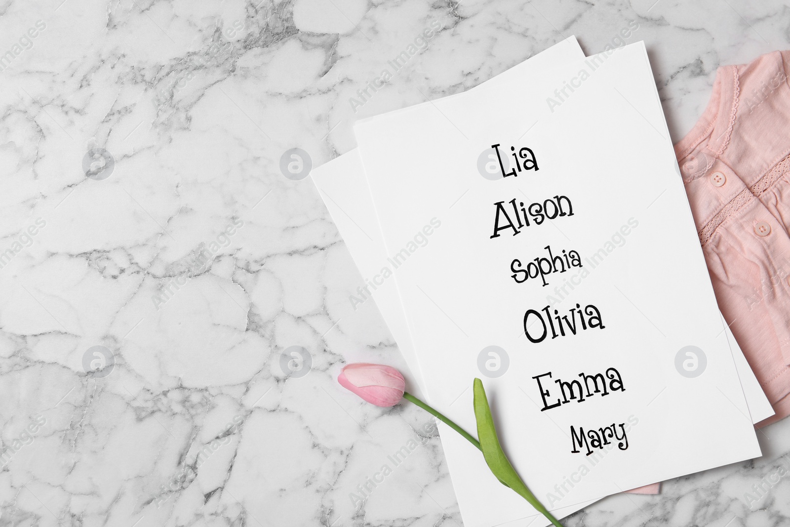 Photo of List of baby names, tulip flower and child's clothes on white marble background, flat lay. Space for text