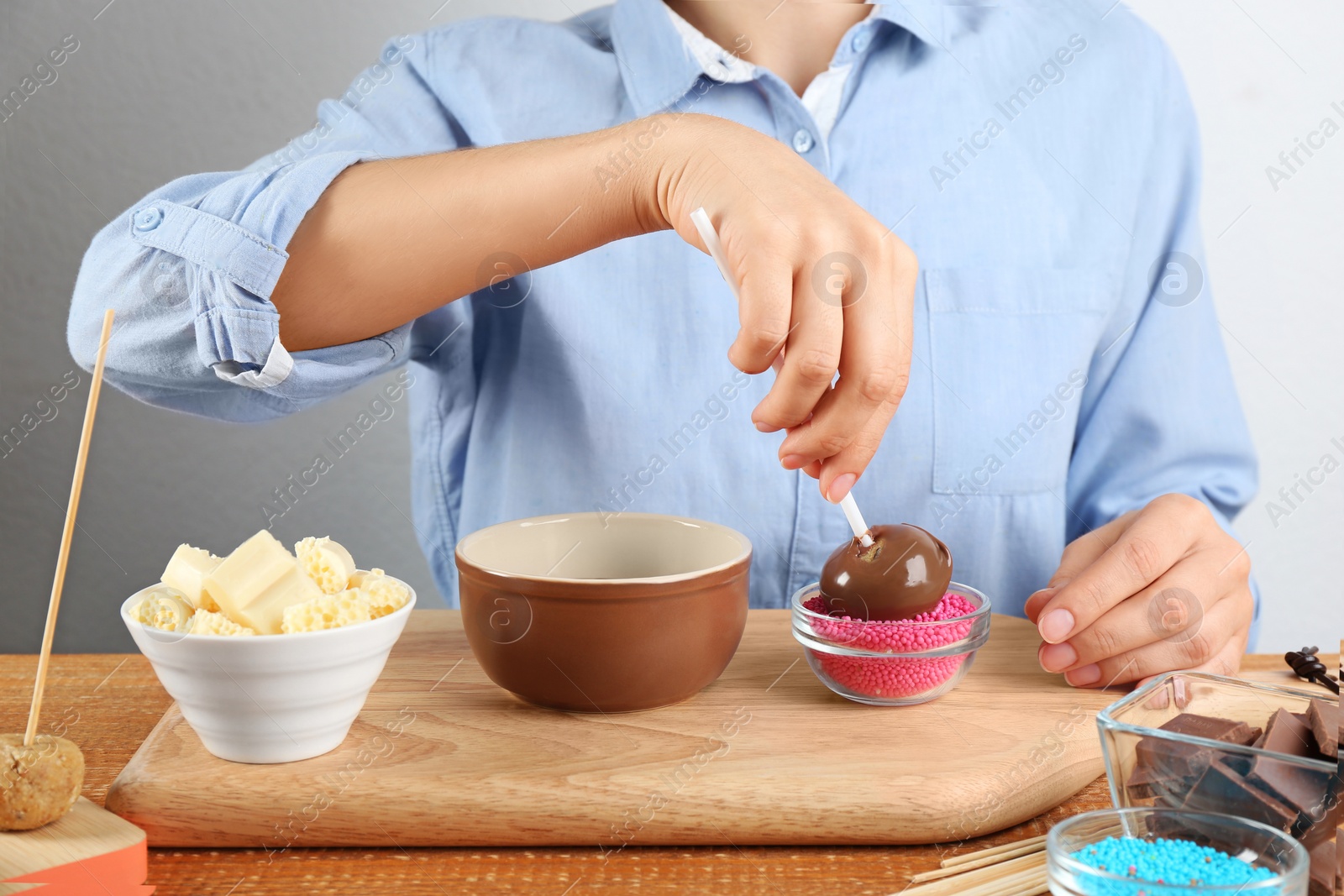 Photo of Young woman putting cake pop into pink sprinkles at wooden table, closeup