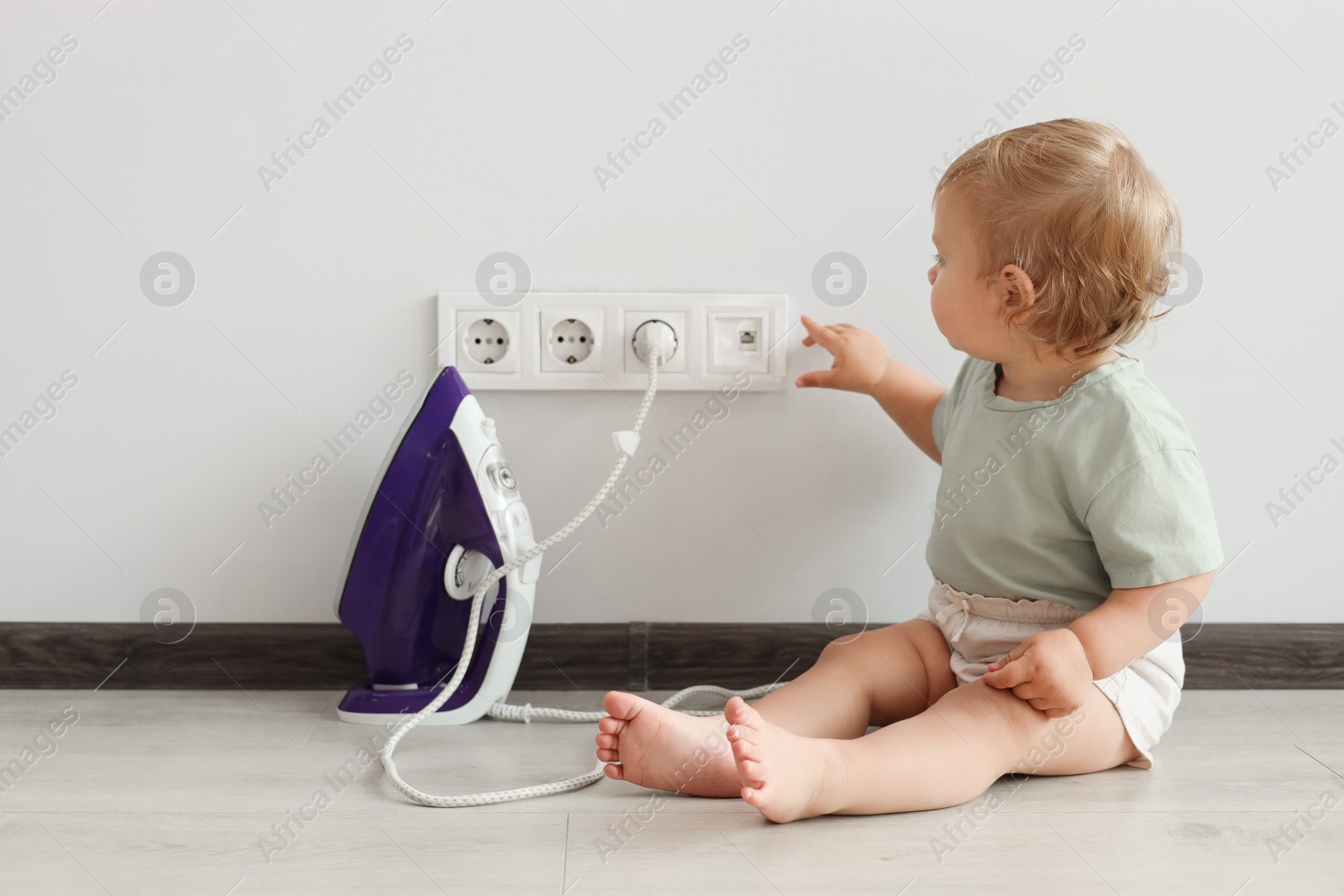 Photo of Cute baby playing with electrical socket and iron plug at home. Dangerous situation