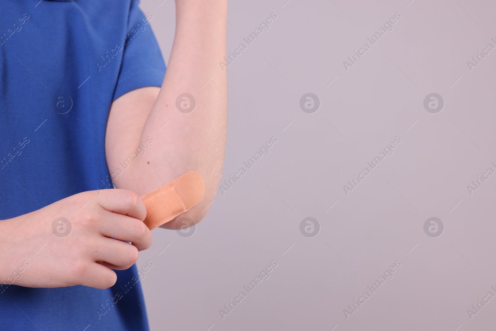 Photo of Little boy putting sticking plaster onto elbow against light grey background, closeup. Space for text