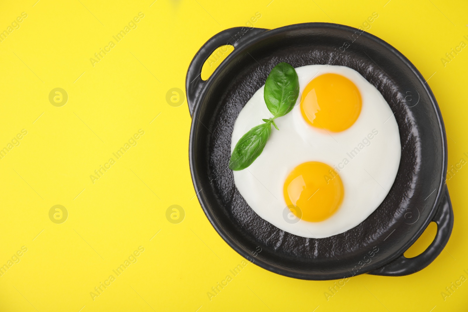 Photo of Tasty fried eggs with basil in pan on yellow background, top view. Space for text