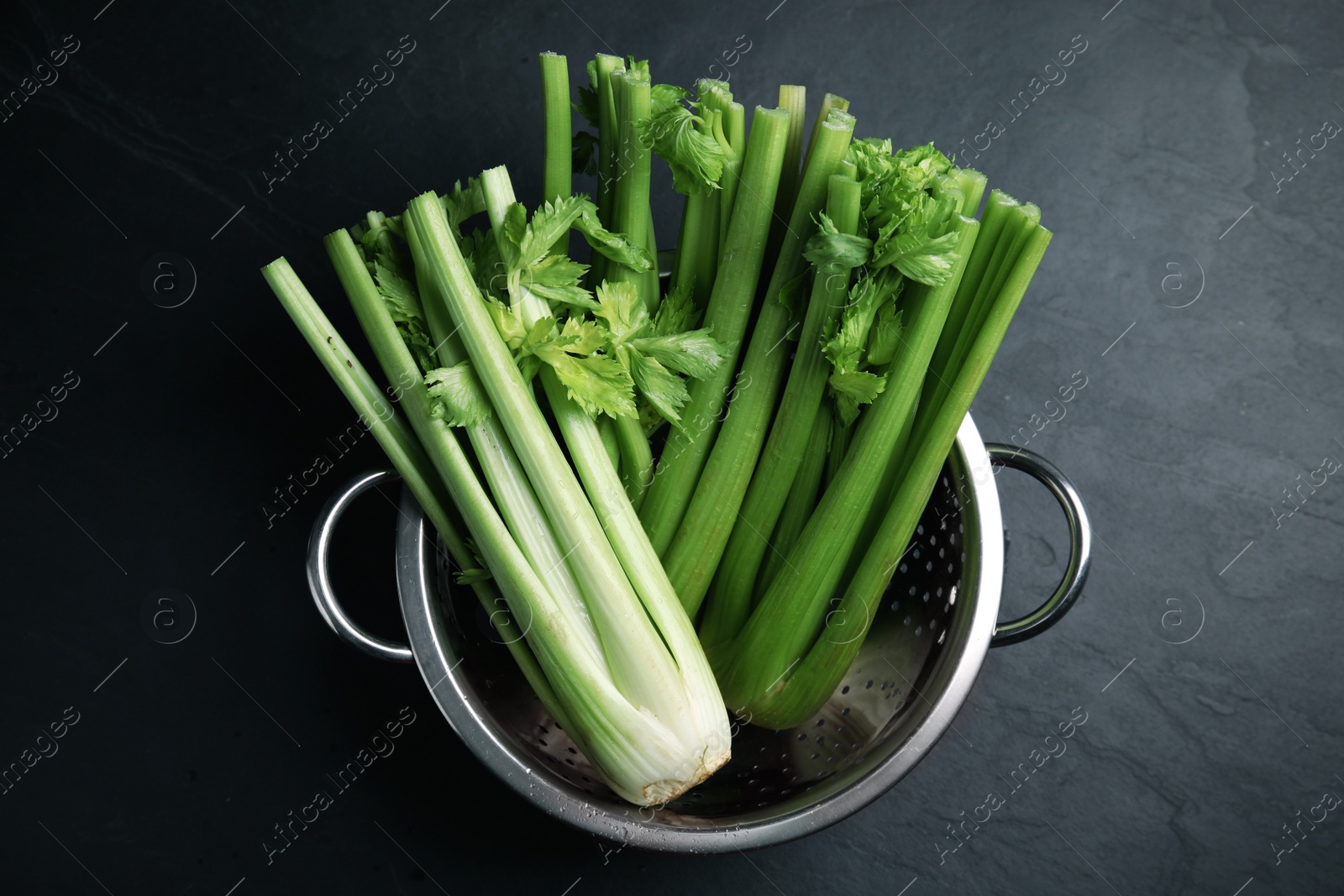 Photo of Fresh green celery in colander on black table, top view