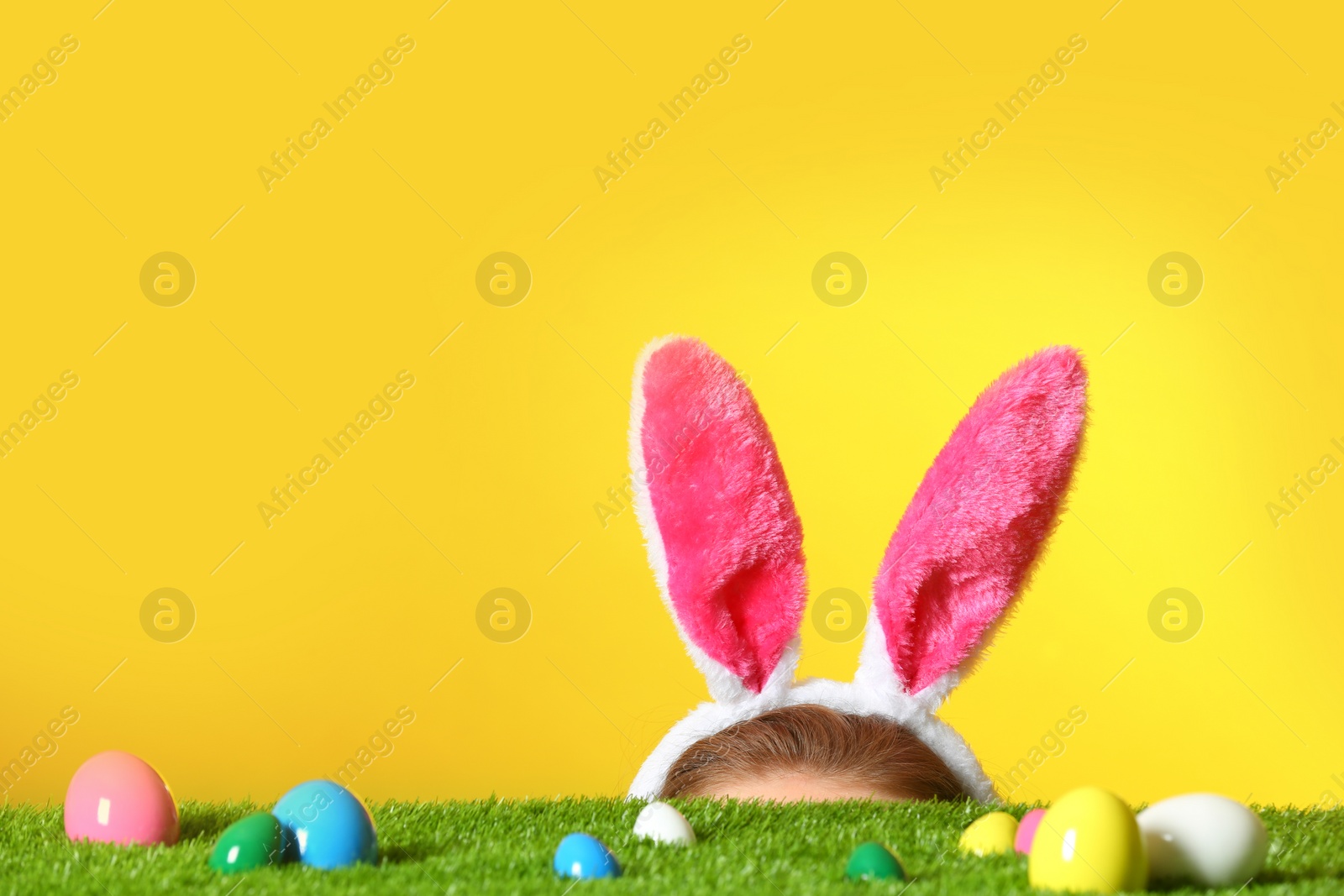 Photo of Easter eggs on green lawn and little girl wearing headband with bunny ears against color background, space for text