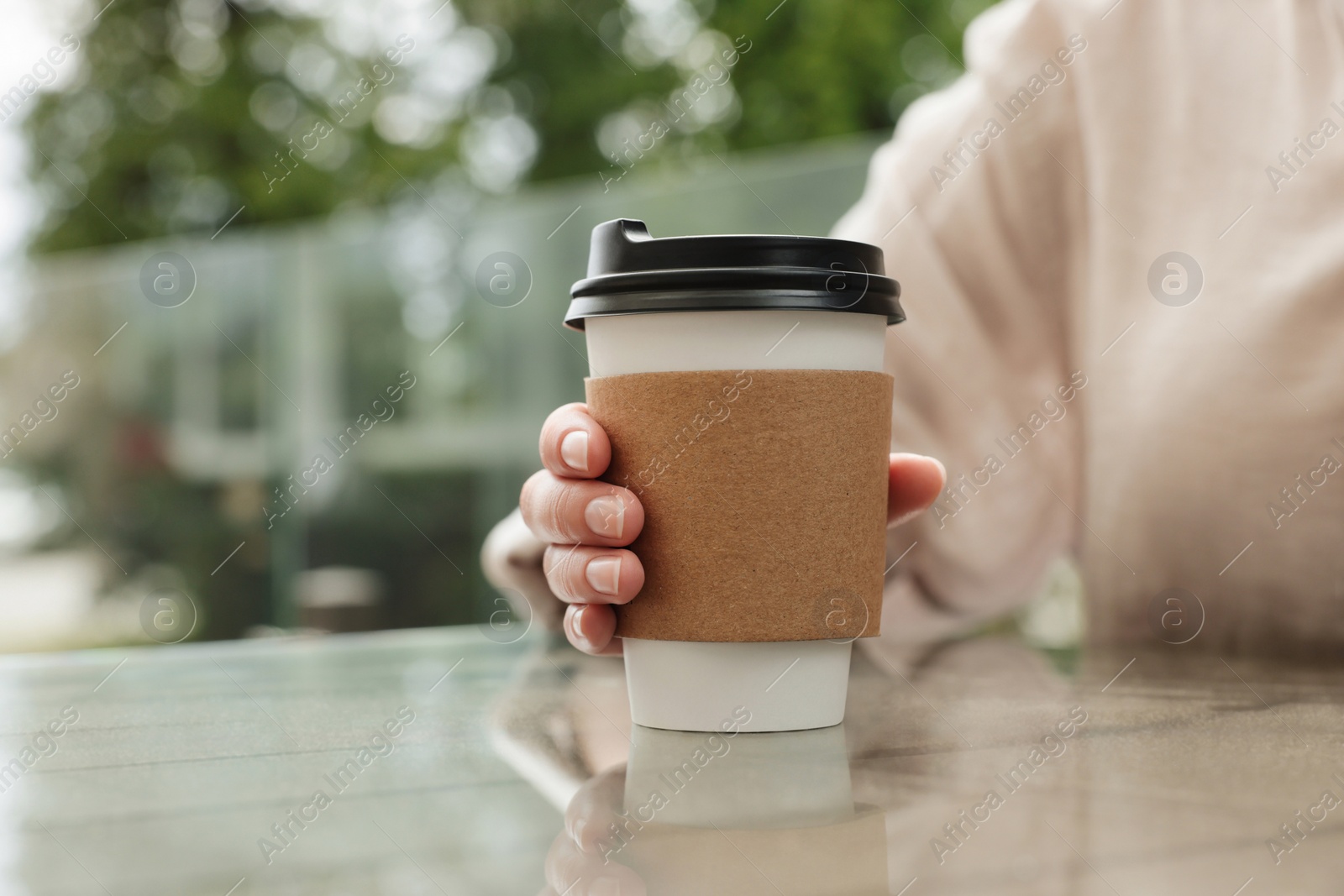 Photo of Woman holding paper takeaway cup at glass table outdoors, closeup. Coffee to go