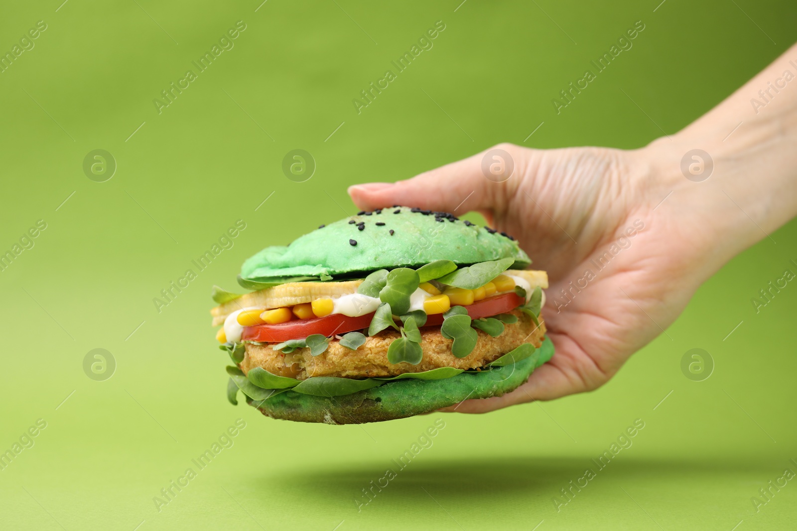 Photo of Woman holding tasty vegan burger with vegetables, patty and microgreens on green background, closeup