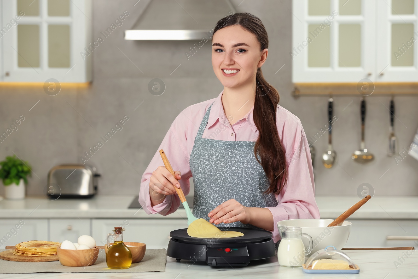 Photo of Happy woman cooking delicious crepe on electric maker at white marble table in kitchen