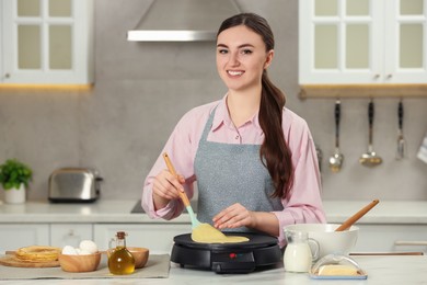 Happy woman cooking delicious crepe on electric maker at white marble table in kitchen