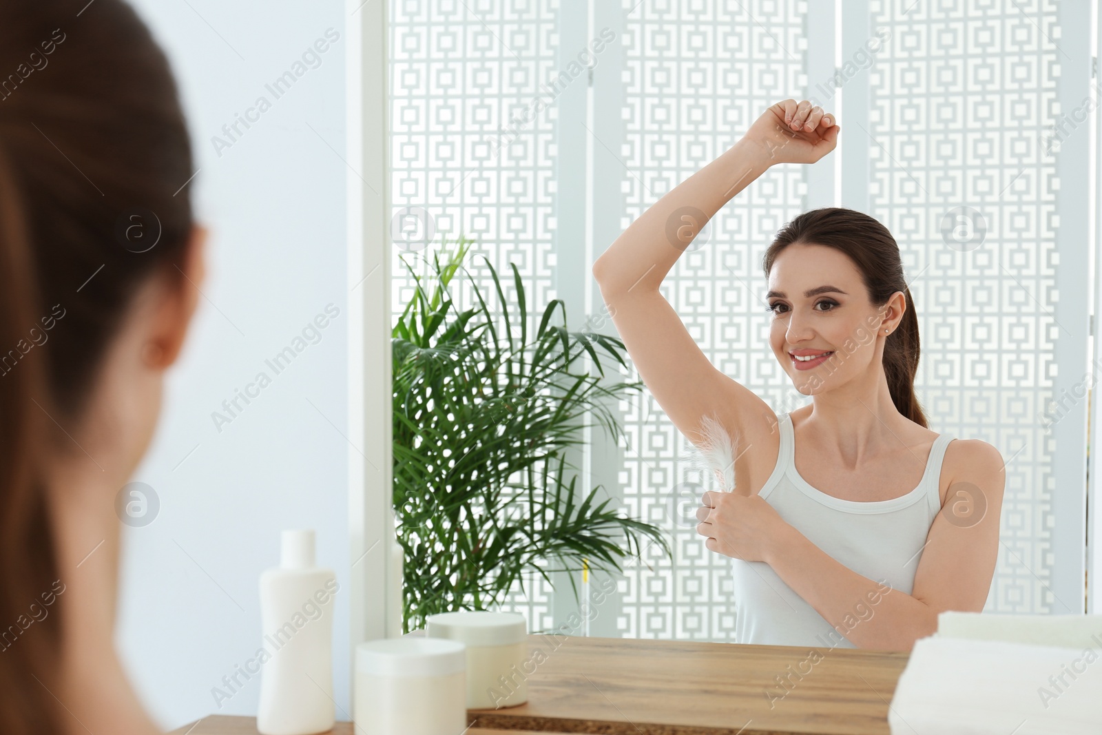 Photo of Young woman touching armpit with feather near mirror at home. Epilation procedure
