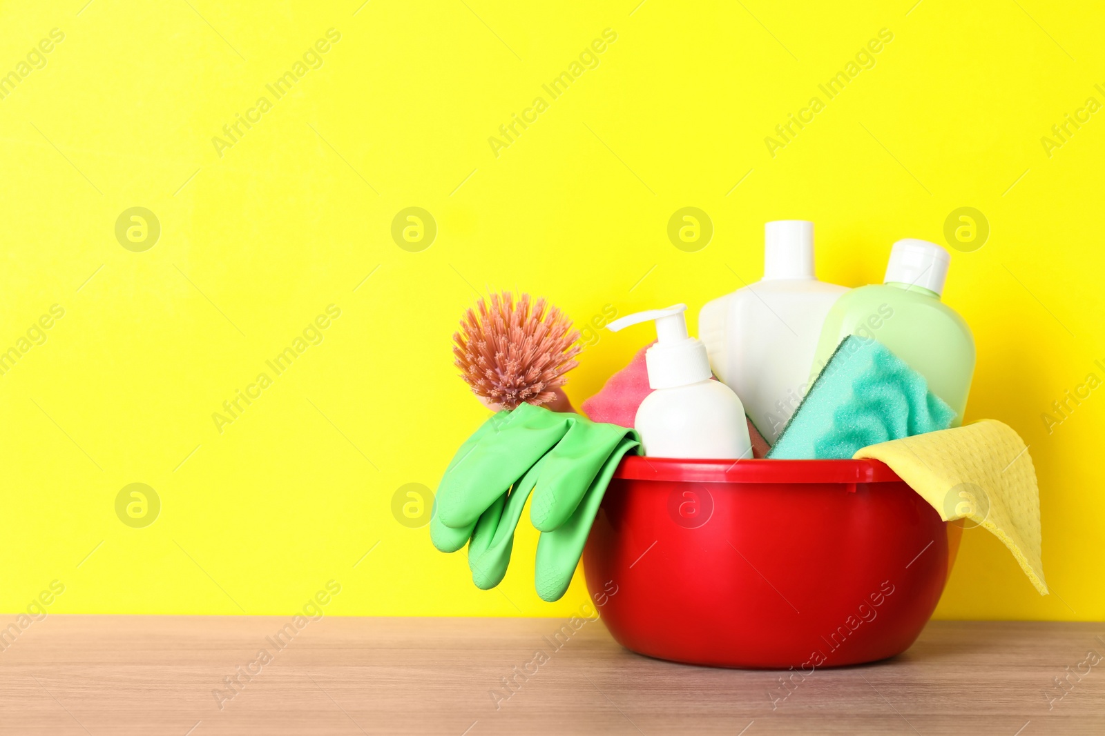 Photo of Basin with different cleaning supplies on wooden floor near yellow wall. Space for text