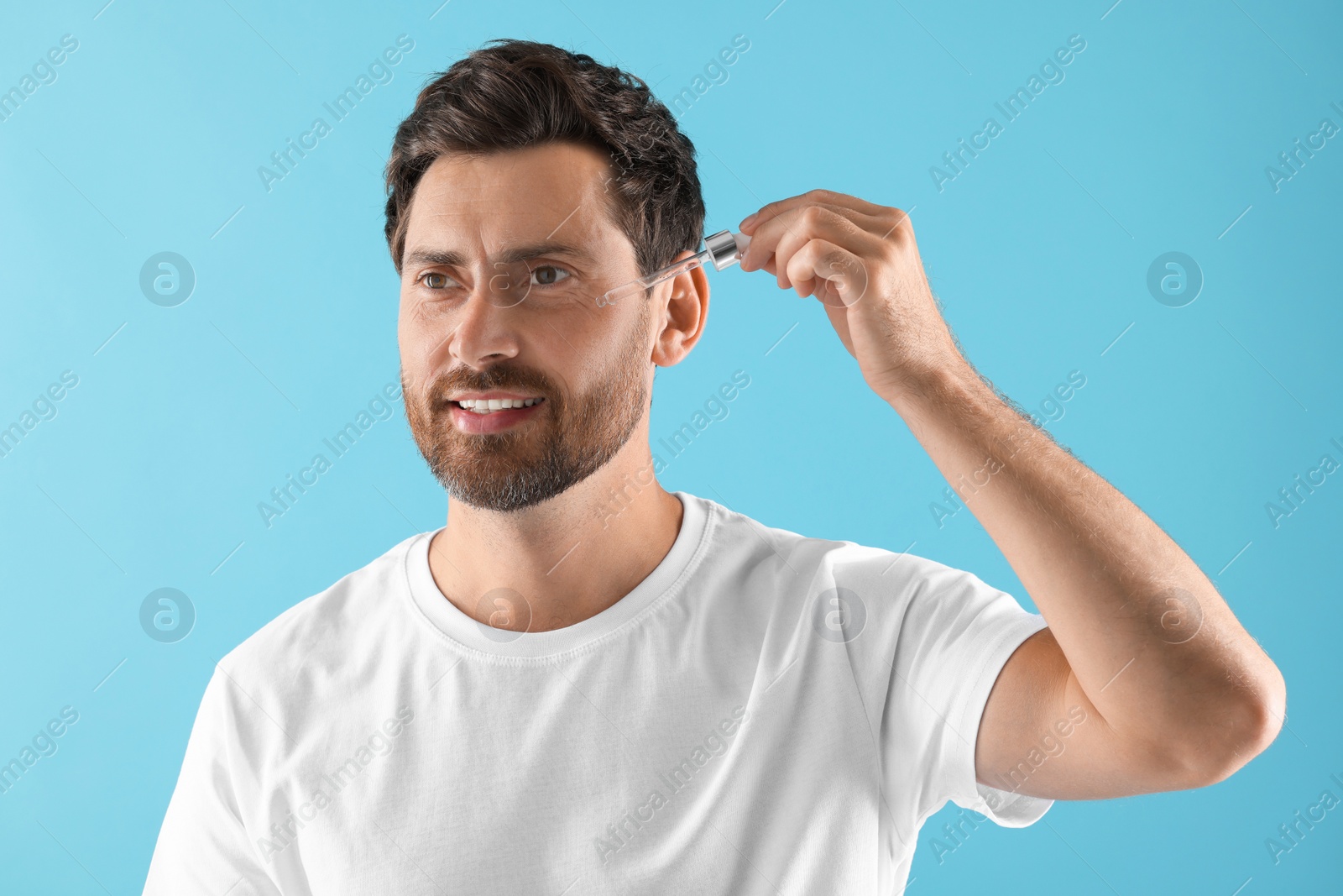 Photo of Smiling man applying cosmetic serum onto his face on light blue background