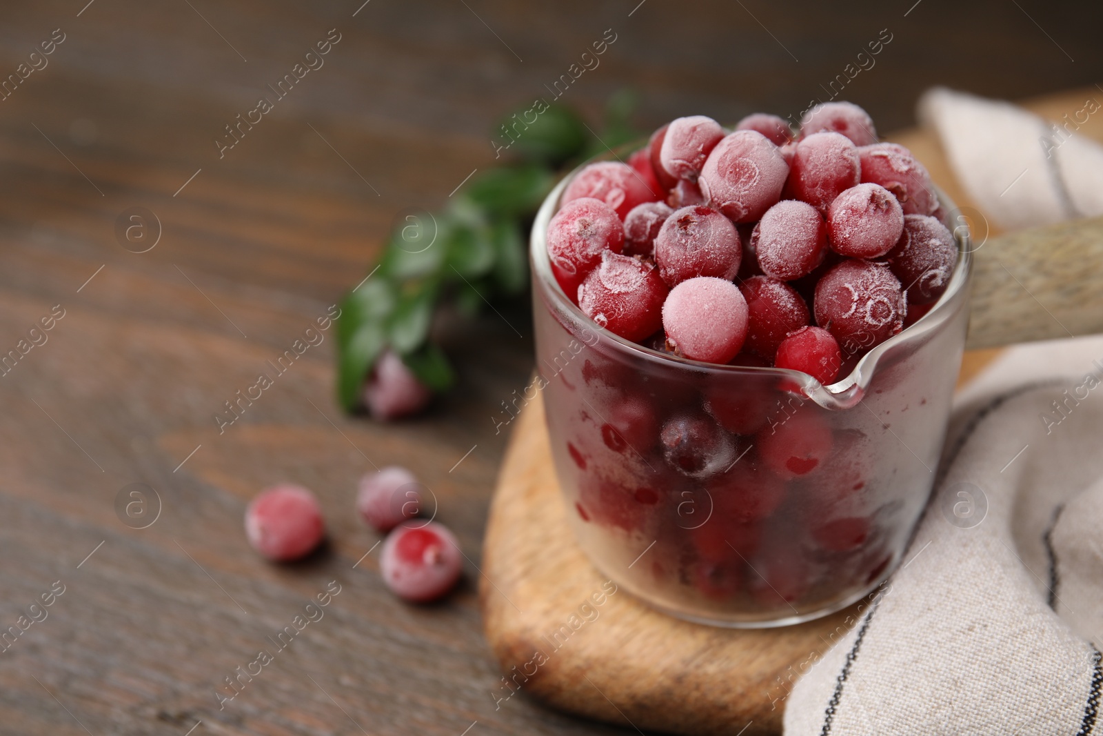 Photo of Frozen red cranberries in glass pot and green leaves on wooden table, closeup. Space for text