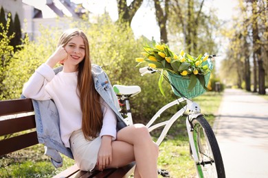 Photo of Beautiful teenage girl sitting on wooden bench near bicycle with bouquet of yellow tulips outdoors. Spring day