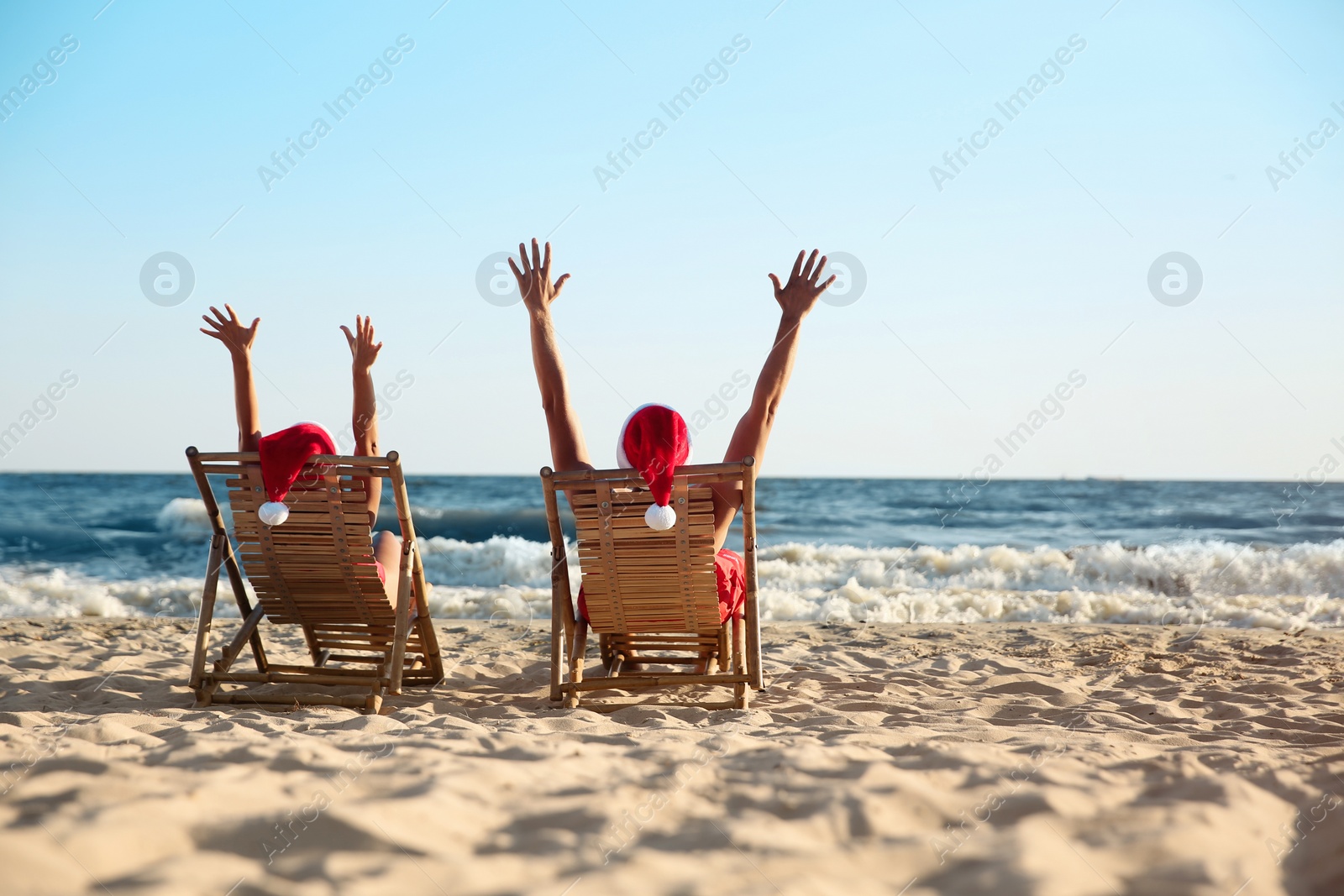 Photo of Lovely couple with Santa hats relaxing on deck chairs at beach, back view. Christmas vacation