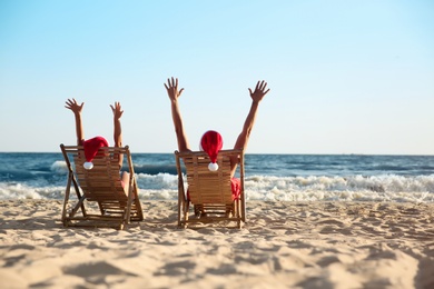 Lovely couple with Santa hats relaxing on deck chairs at beach, back view. Christmas vacation