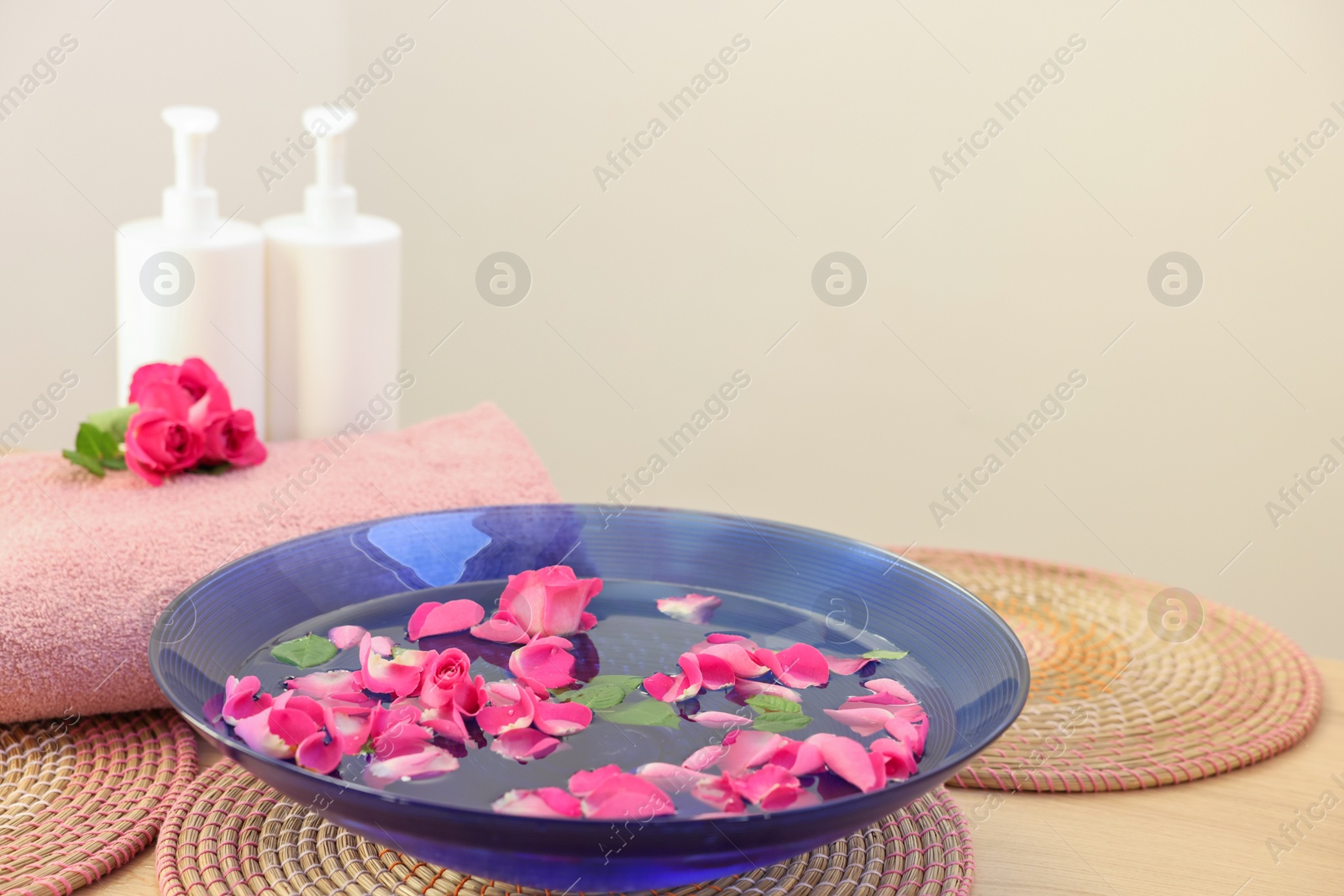 Photo of Pink roses, petals in bowl with water, cosmetic products and towel on wooden table. Space for text