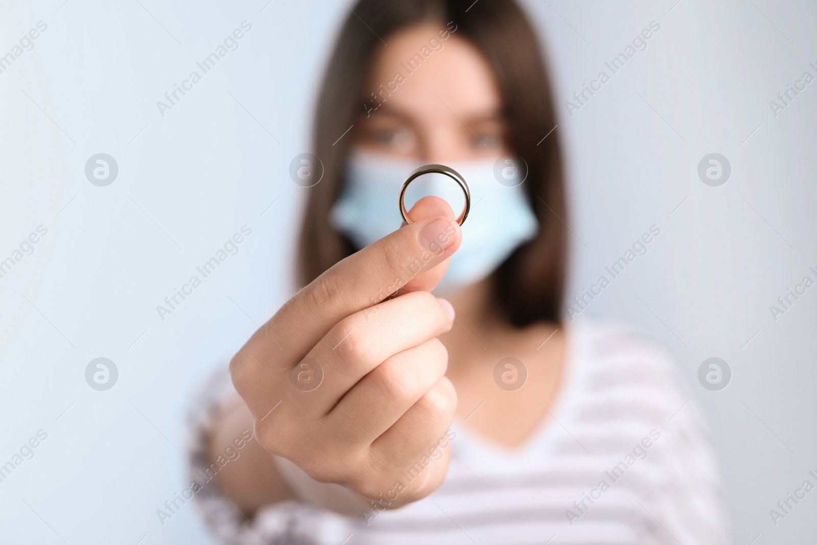 Photo of Woman in protective mask holding wedding ring against light background, focus on hand. Divorce during coronavirus quarantine