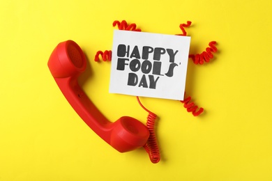 Photo of Telephone handset and Happy Fools' Day note on yellow background, flat lay