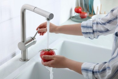 Photo of Woman washing fresh ripe tomato under tap water in kitchen, closeup