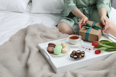 Photo of Tasty breakfast served in bed. Woman with gift box, desserts, tea, flowers and I Love You card at home, closeup