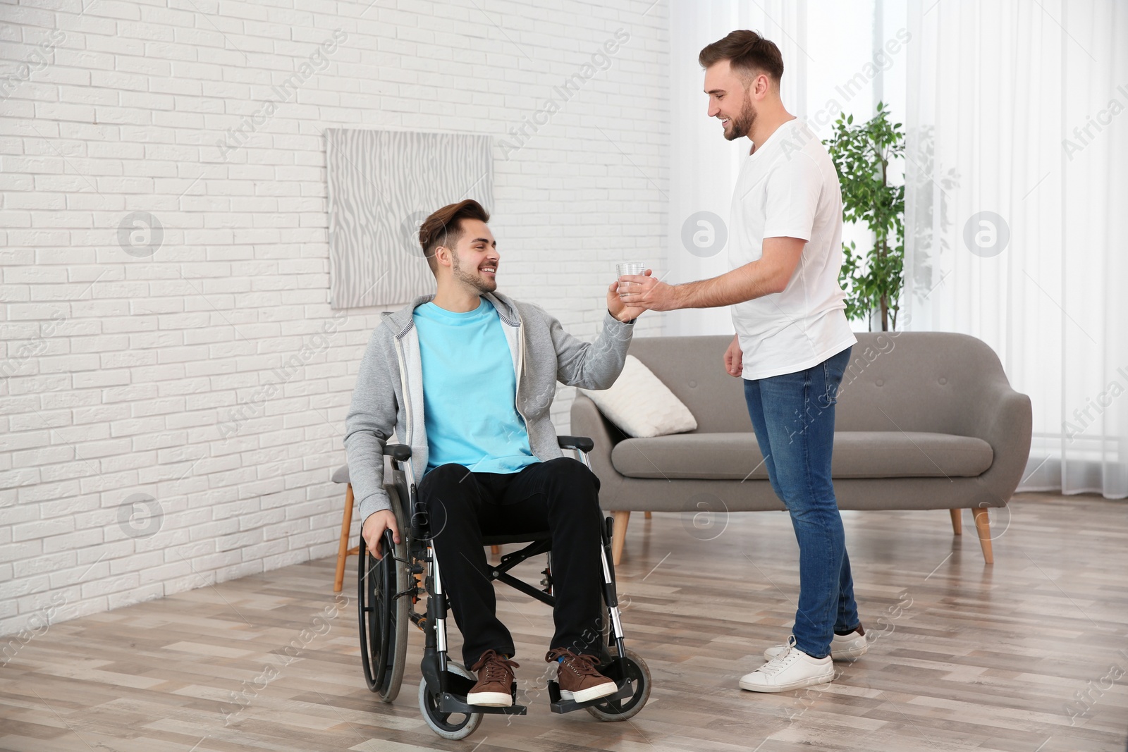 Photo of Volunteer giving glass of water to man in wheelchair at home. Helping disabled