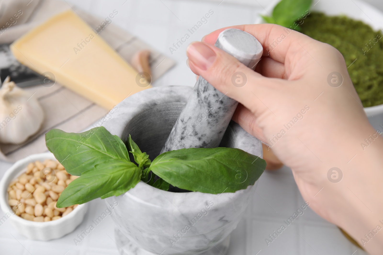 Photo of Woman mashing basil for pesto sauce in mortar at white table, closeup