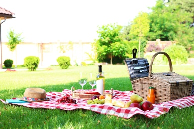 Picnic basket with products and bottle of wine on checkered blanket in garden