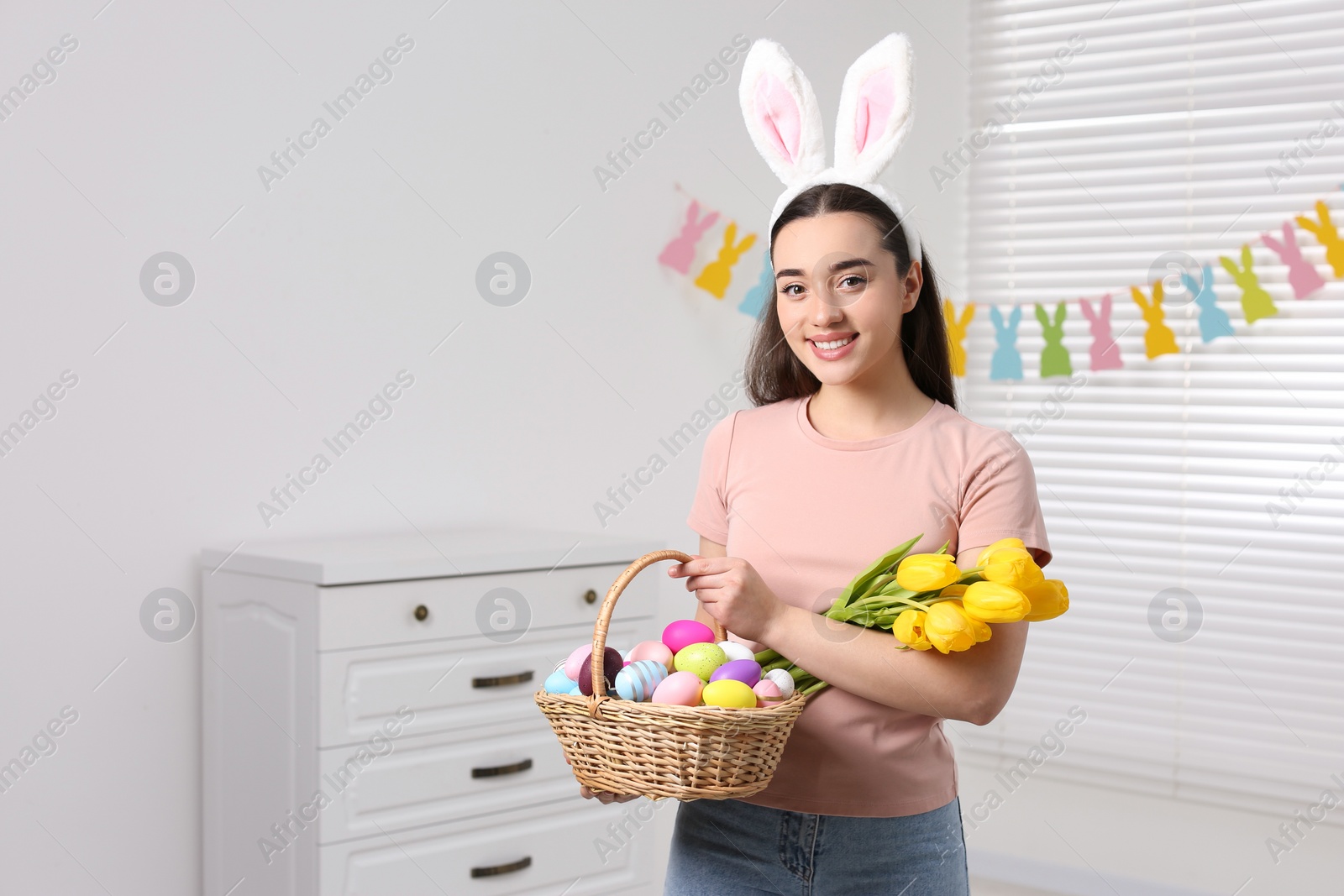 Photo of Happy woman in bunny ears headband holding wicker basket with painted Easter eggs and bouquet of flowers indoors. Space for text