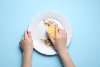 Woman washing dirty plate with sponge on light blue background, top view