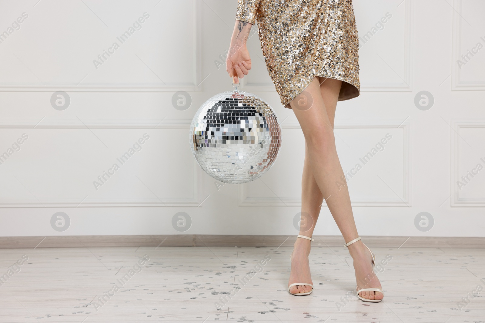 Photo of Woman in high heeled shoes and golden dress with disco ball near white wall indoors, closeup