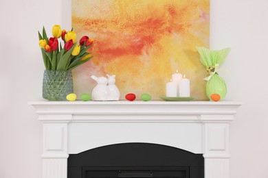 Photo of Easter decorations. Bouquet of tulips in vase, burning candles and bunny figures on fireplace at home