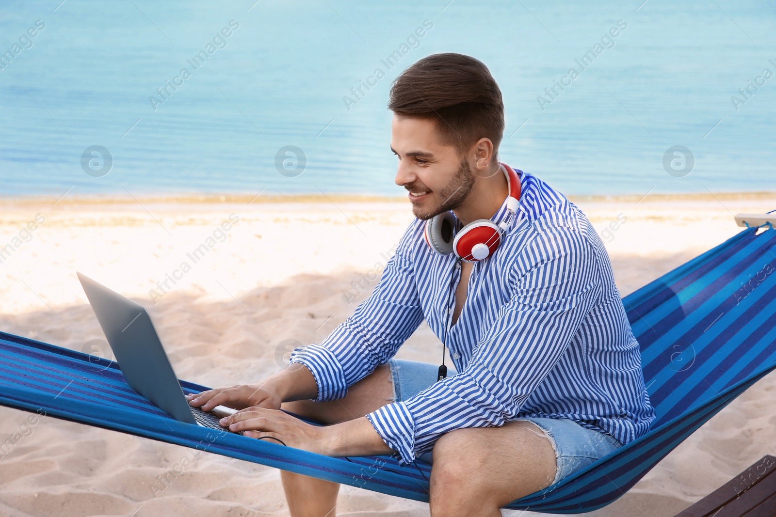 Photo of Young man with laptop and headphones sitting in hammock at seaside