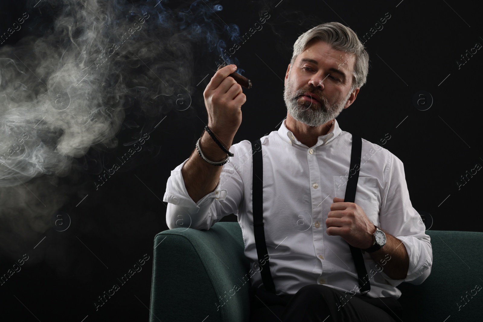 Photo of Bearded man smoking cigar on sofa against black background. Space for text