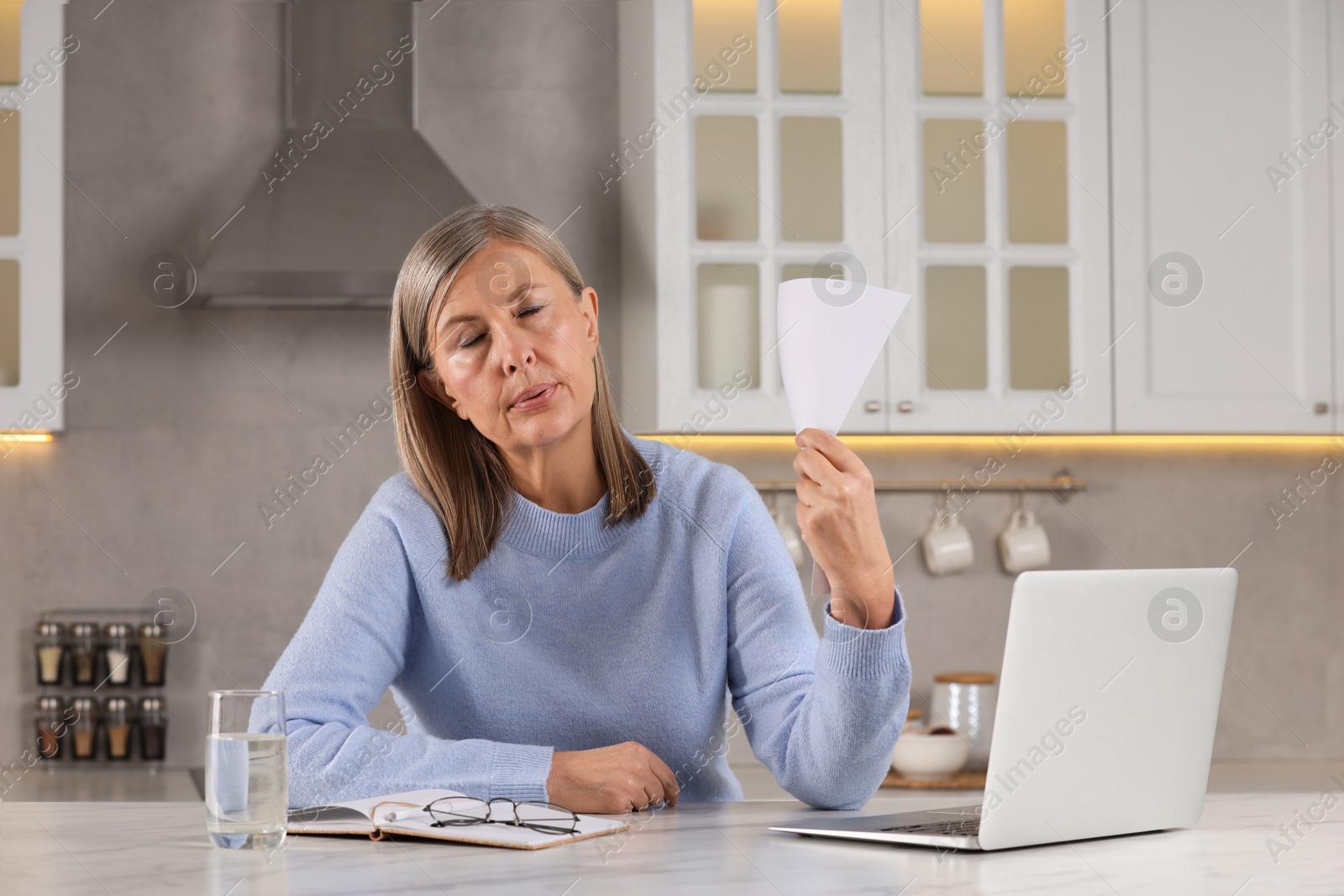 Photo of Menopause. Woman waving paper to cool herself during hot flash at table in kitchen