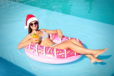 Young woman wearing Santa Claus hat with refreshing drink on inflatable ring in swimming pool. Christmas vacation