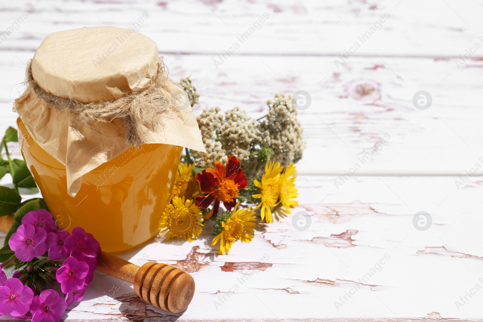 Photo of Glass jar of honey, dipper and different flowers on white wooden table. Space for text