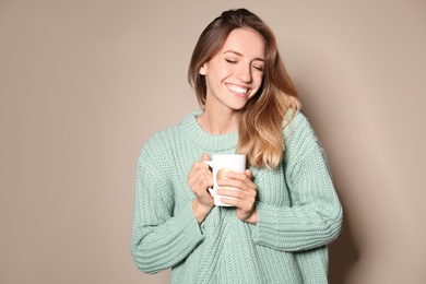 Image of Happy young woman in warm sweater with cup of hot drink on beige background