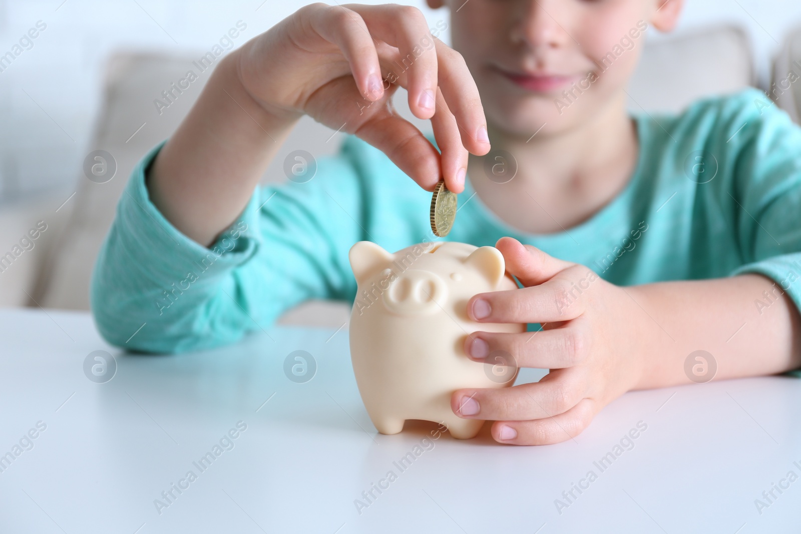 Photo of Little boy putting coin into piggy bank at white table indoors, closeup
